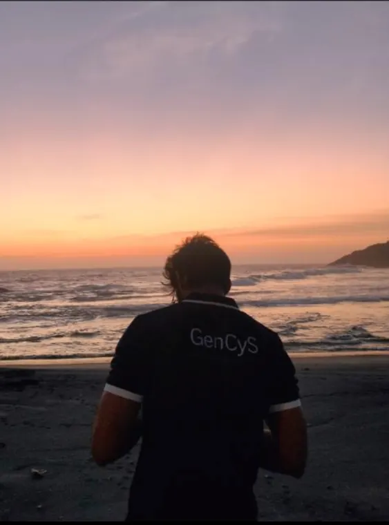 A person in a GenCys shirt looks out over the ocean at sunset.